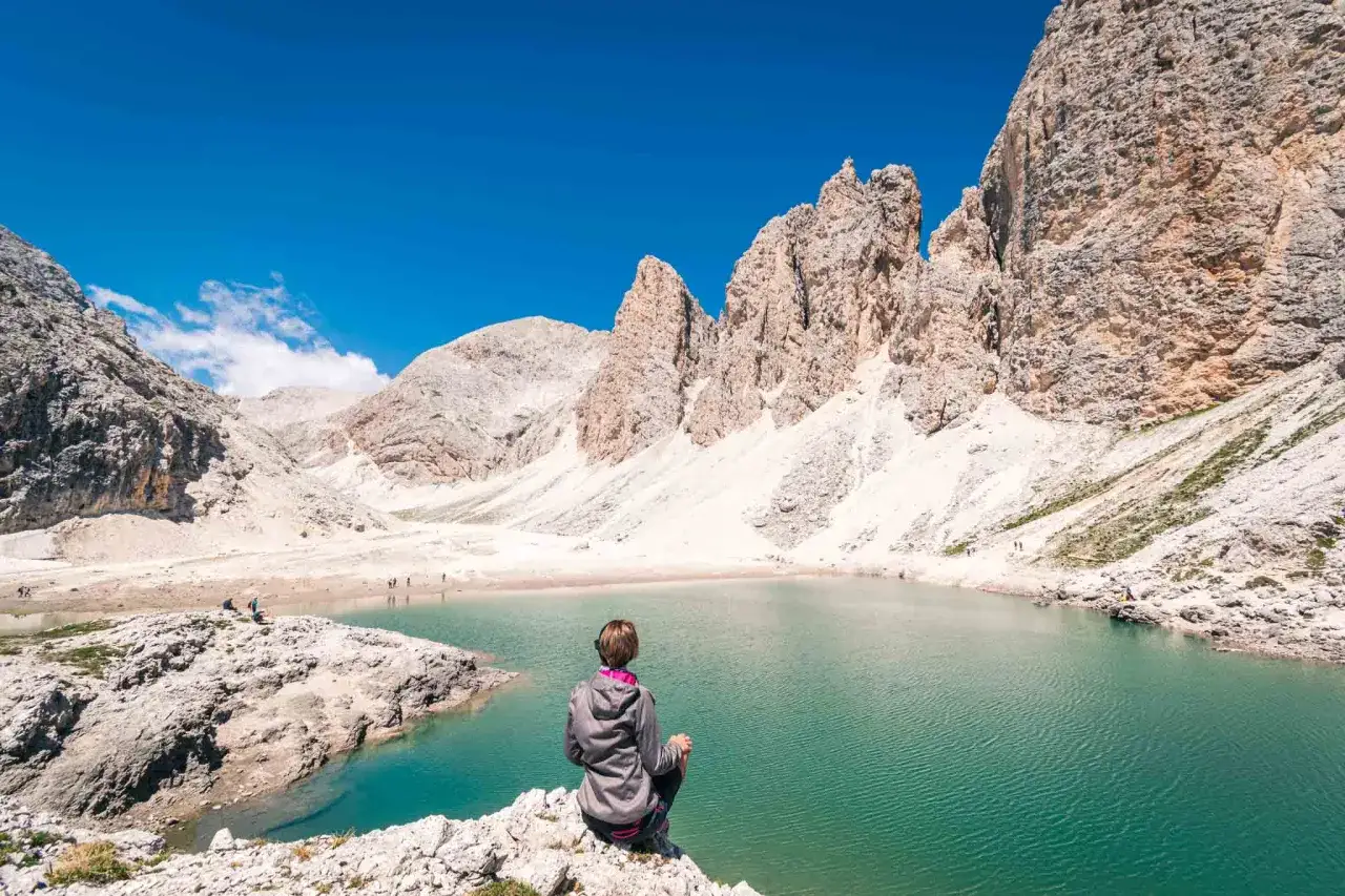 Donna seduta ammira un lago alpino turchese, circondato da imponenti vette rocciose. Un'immagine che cattura la bellezza delle val di fassa escursioni pi&ugrave; belle.