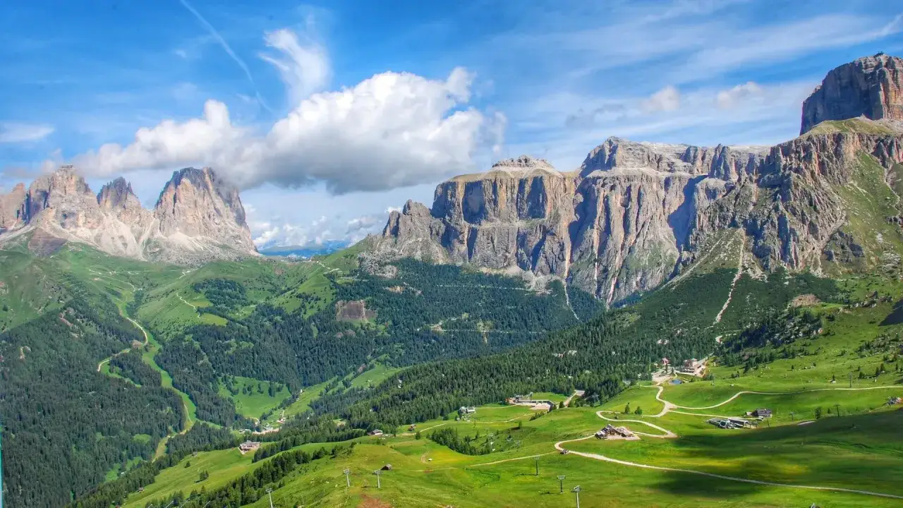 Viste mozzafiato delle Dolomiti Trentino Alto Adige, con valli verdi, boschi fitti e imponenti pareti rocciose sotto un cielo azzurro con nuvole bianche.
