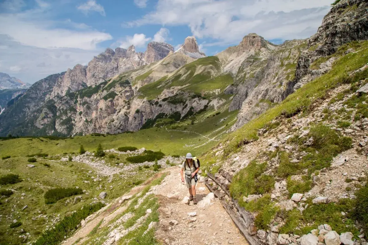 Un escursionista percorre un sentiero nelle Dolomiti facili, con imponenti vette rocciose e verdi pendii sullo sfondo.