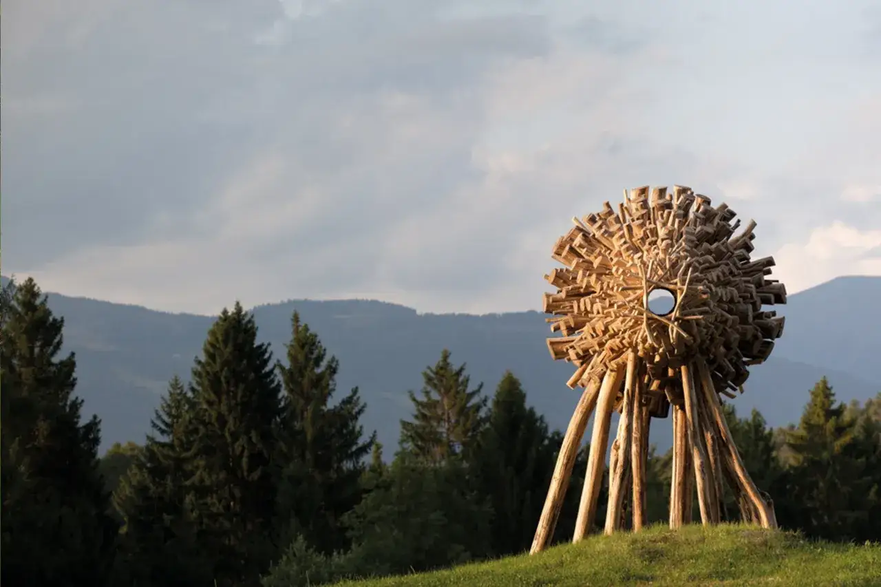 Installazione artistica in legno a Borgo Valsugana, Arte Sella. Struttura sferica su gambe, circondata da alberi e montagne.