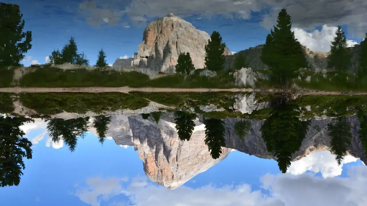 Riflesso speculare di un maestoso itinerario Dolomiti: montagne rocciose, verdi abeti e cielo azzurro con nuvole bianche.