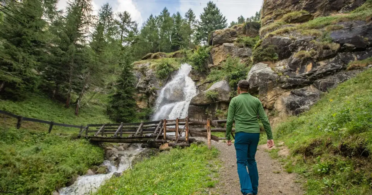 Un escursionista ammira una cascata nel Val di Sole in estate, con un ponte di legno che attraversa il torrente.
