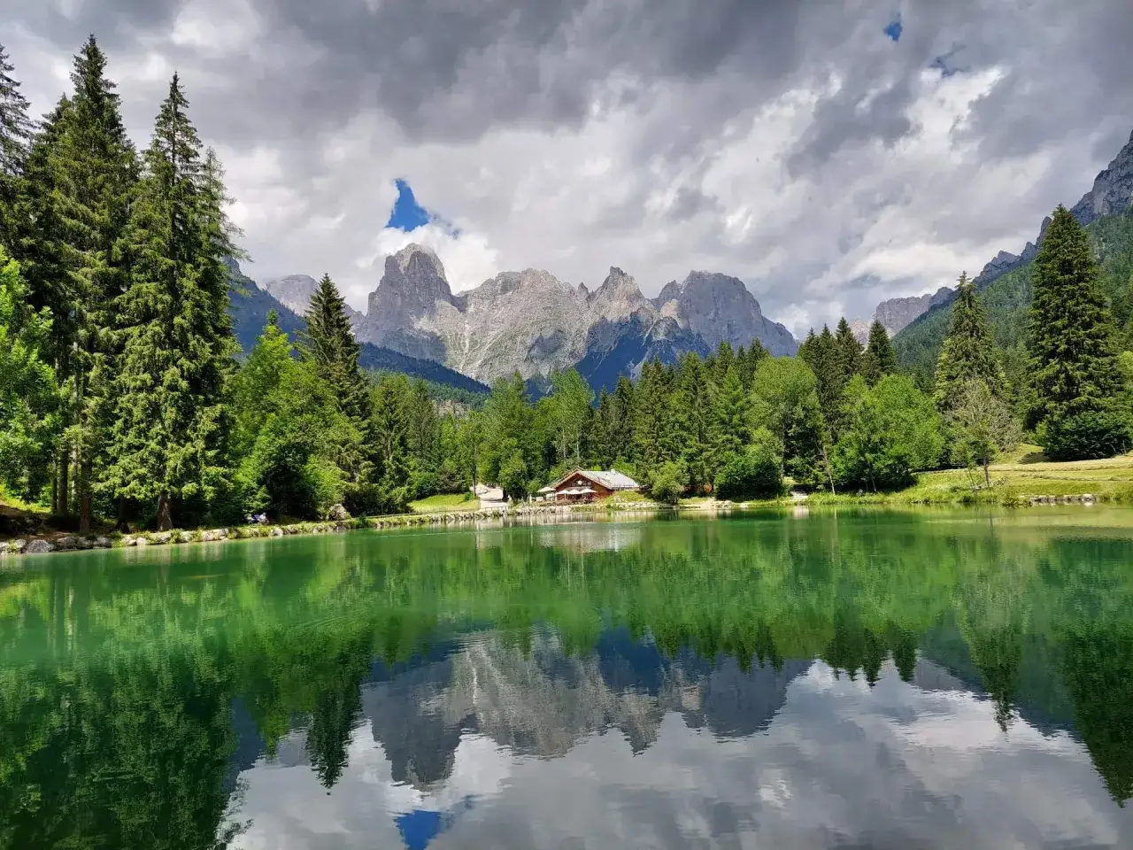 Riflessi alpini sul lago di Passo Cereda, con un rifugio tra i boschi e montagne imponenti.