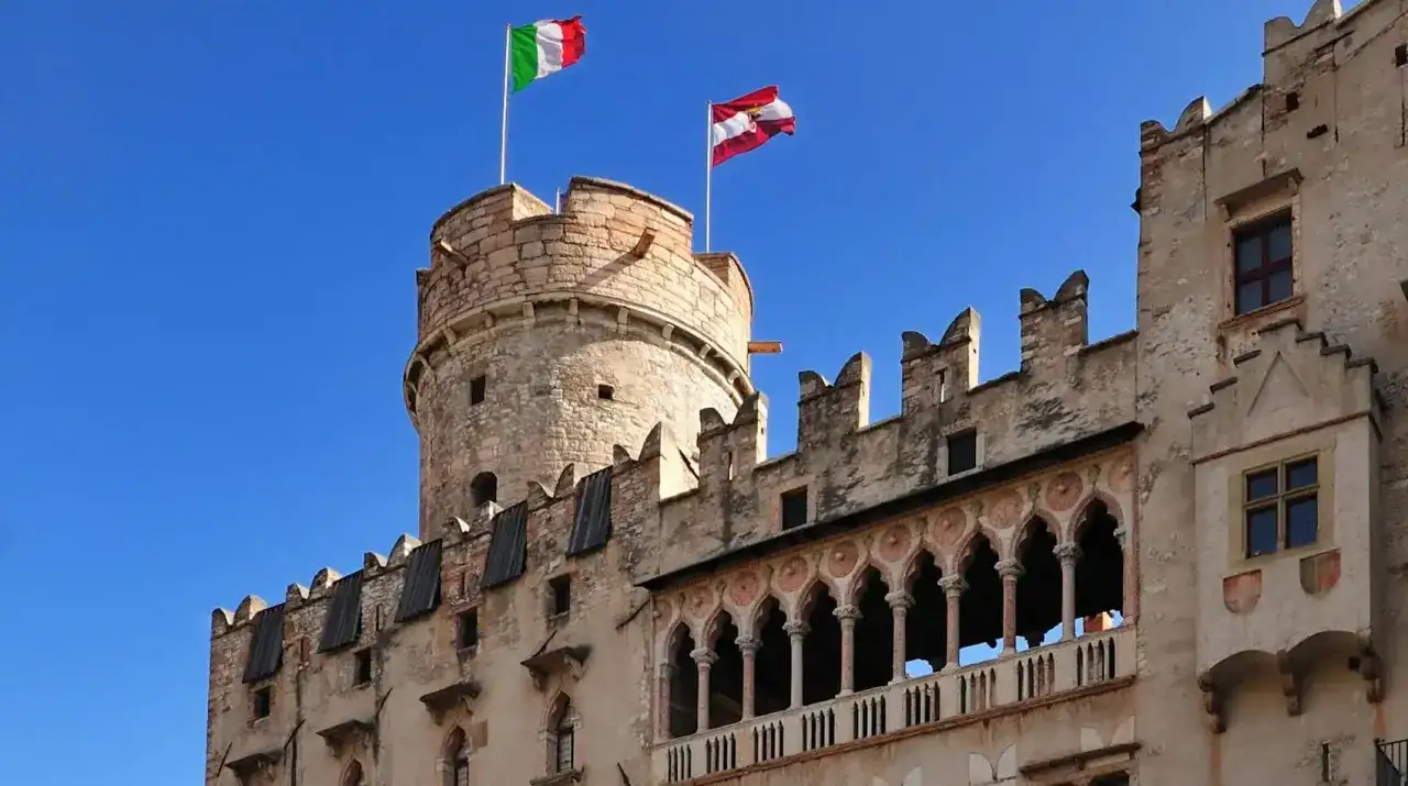 Il castello del Buonconsiglio domina il cielo azzurro con le sue torri e mura antiche, sventolando le bandiere italiana e austriaca.