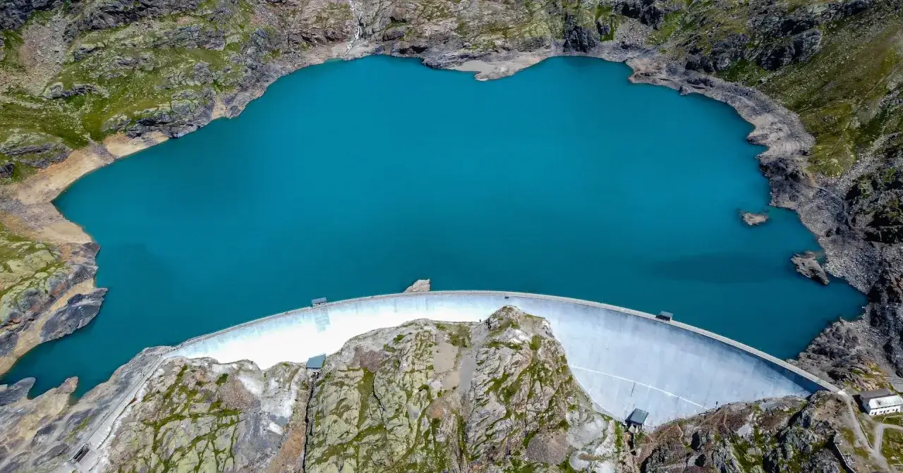 Vista aerea del lago del Careser, un bacino artificiale alpino con acque turchesi circondato da rocce e vegetazione. La diga curva domina la scena.