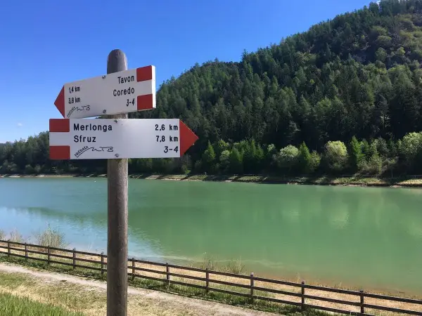 Cartello con indicazioni per Tavon di Coredo, Merlonga e Sfruz, con lago verde e bosco sullo sfondo.