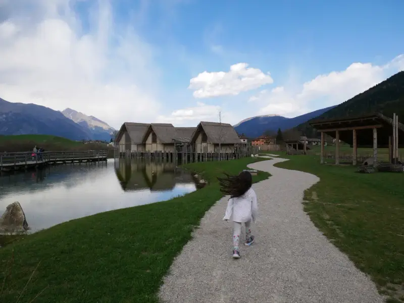 Bambina corre su sentiero ghiaioso verso villaggio palafitticolo nelle Alpi Giudicarie. Montagne sullo sfondo.