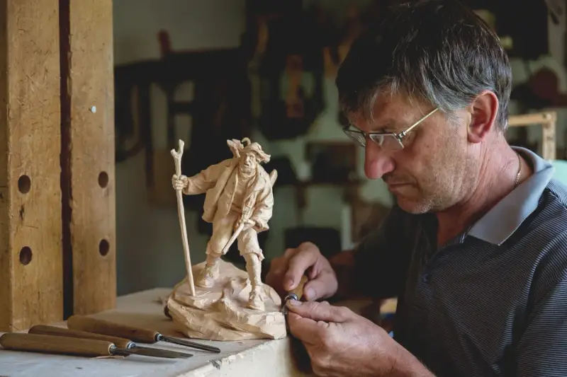 Maestro intaglia un pastore con bastone, un esempio di artigianato legno trentino.