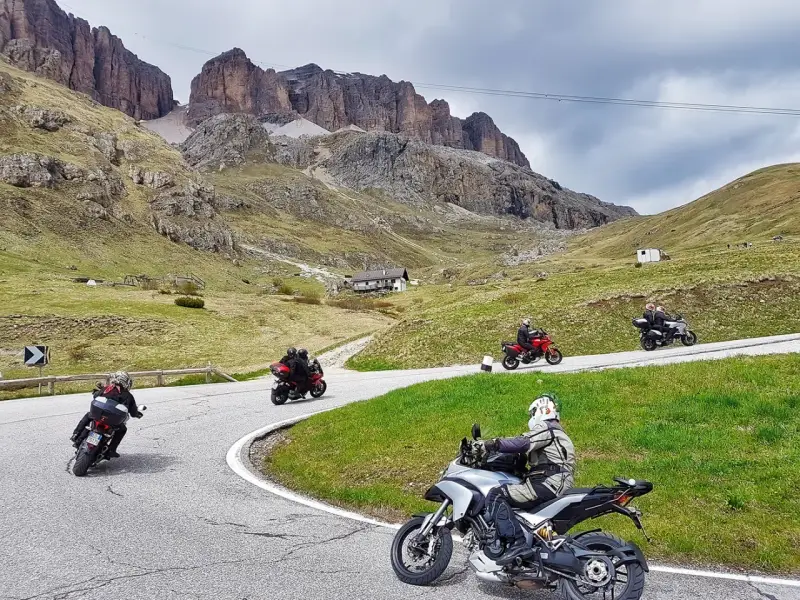 Motociclisti affrontano tornanti sui **Dolomiti passi**, con imponenti vette sullo sfondo.