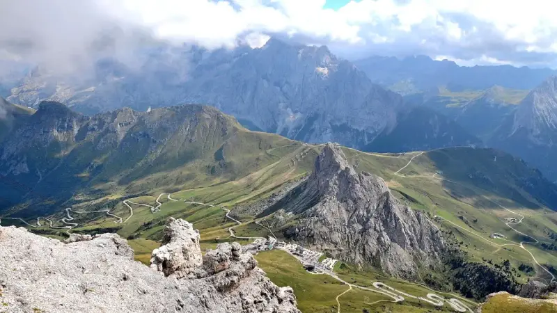 Panoramica mozzafiato del **Passo Pordoi** con strade tortuose che si snodano tra verdi pendii e imponenti vette rocciose, sotto un cielo nuvoloso.