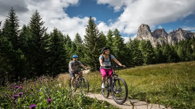 Coppia in mountain bike su sentiero a Passo Campolongo, con bosco di abeti e montagne sullo sfondo.