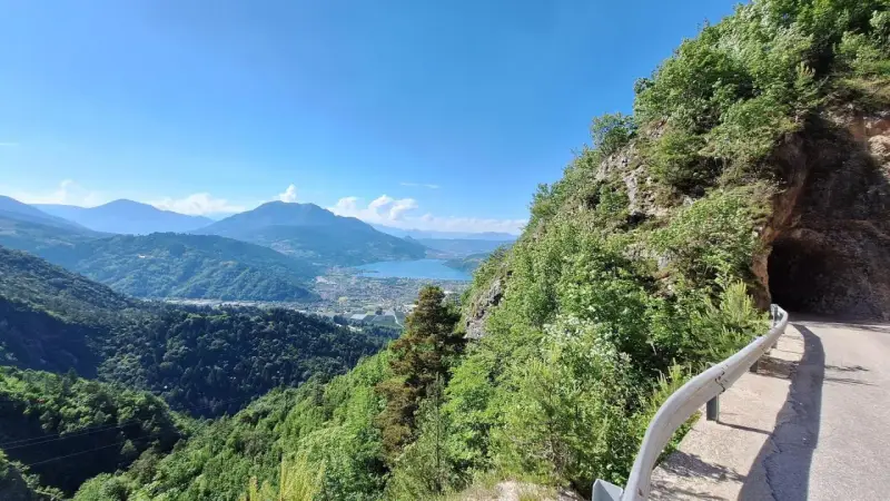 Vista panoramica dal Monte Vezzena con lago azzurro e montagne sullo sfondo. Strada con galleria scavata nella roccia.